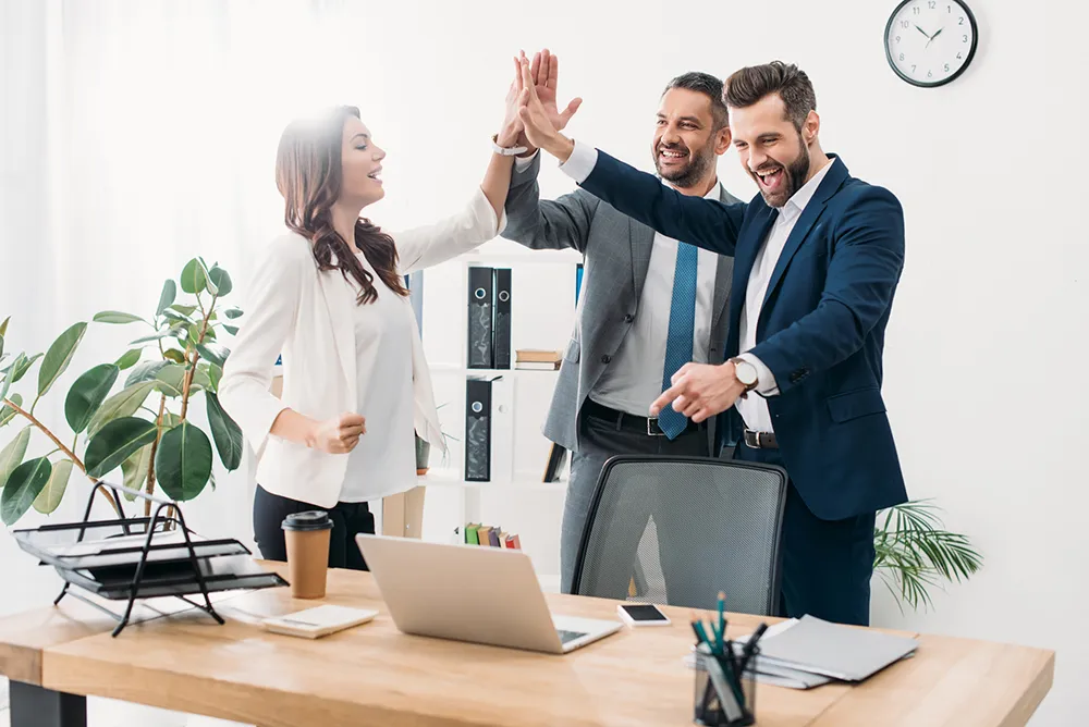 Colleagues standing near table with laptop and highing five in office